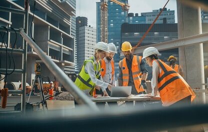 construction site with a crew of people in hardhats and reflective vests around a table looking at plans on a laptop